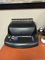 Front view of black GBC CombBind C210E document binding machine on wooden desk with small speakers and a small plant nearby.