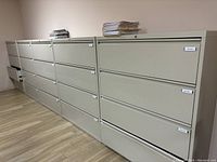 Side view of a row of metal filing cabinets in light beige color showing multiple cabinets with three drawers each in an office setting on wood floor.