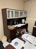Office credenza shown with clutter and papers on surface, dark wood with frosted glass cabinets on upper section.