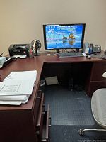 Small dark wood office desk with computer equipment and office accessories on top. Desk drawers partially open, chair on casters, and floor mat visible.