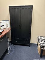 Tall black wood filing cabinet with double doors and a bottom drawer, set against a beige wall with a desk nearby.