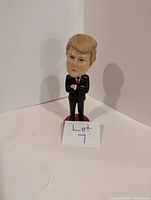 Front view of the Donald Trump bobblehead figure shows the figure with arms crossed, detailed facial features, and hair sculpted in a comb-over style, standing on a red base.