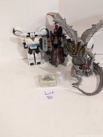 Photo showing two robot figures, one white and blue and one black and red, and a detailed dragon figurine with wings. Also visible is a small sealed pack of cards or accessories in front.