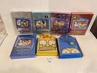 Seven boxed sets of The Simpsons DVDs displayed upright, showing front covers with various Simpsons characters and season labels, including the 2007 Collector's Edition for season 1.