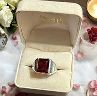 Front angle of the men's sterling silver ring with a rectangular red stone set in center inside beige velvet-lined PEOPLES box surrounded by flowers and candlelight.