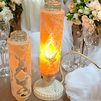 Lamp with lit peach-toned floral etched glass shade on Bakelite base and extra similar shade beside it.