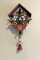 Front view of the clock showing the clock face, chalet-style wooden housing, figurines, and pinecone weights.