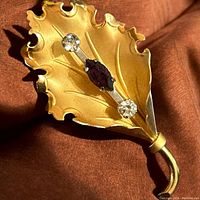 Close-up photo of the leaf-shaped brooch highlighting gold tone finish, scalloped edges, silver bar with one marquise red gemstone and two clear round gemstones.