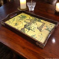 Photo of the vintage wooden Chinese tea tray with brass corner corners, inset traditional artwork panel showing figures in a garden scene. Tray is placed on wooden surface with candle and glassware around.