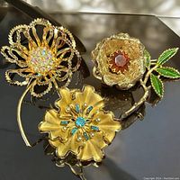 Three decorative vintage flower brooches laid on reflective surface showing details and color of stones and enamel