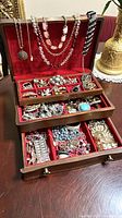 Photo of vintage wooden jewelry box opened showing several necklaces hanging on lid and various rings, bracelets and earrings organized in compartments and drawers with red velvet lining.
