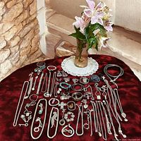 Full display of various vintage silver-toned jewellery, including chains, rings, bracelets, earrings, brooches, and pendants arranged around a flower vase on a burgundy cloth.