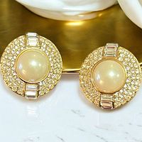 Close-up front view of one earring showing central large faux pearl surrounded by small round and rectangular clear rhinestones set in gold-tone metal.