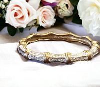 Front and angled views of the gold tone Swarovski bracelet adorned with clear crystals in a bamboo design, shown with floral and wood background.