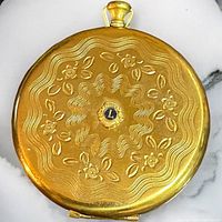 Top view of vintage gold tone compact with embossed floral and wave pattern and a central blue enamel medallion with 'L'