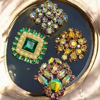 Four vintage rhinestone brooches displayed on a reflective surface, showing colors and design details