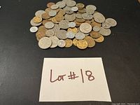 Photo showing a top view of approximately 70 assorted old coins spread out on a black surface with a handwritten lot number 18 on white paper.