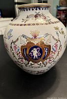 Front view of one vase showing round shape, cherub shield design with floral motifs and gold accents.