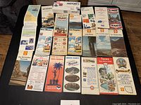 Top-down photo showing a spread of approximately 25 vintage maps and brochures laid out on a wooden surface, variety of regions and gas station brands represented.