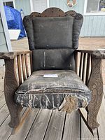 Front view of the vintage wooden rocking chair showing worn black leather seat heavily torn at the front edge, padded backrest, and detailed carved wooden arms and legs.