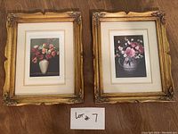 Pair of floral artwork prints in ornate gold frames with cream mats, placed side by side on wooden floor