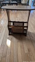 Full front view of the oval wooden side table showing cane magazine rack on one side and wooden legs on hardwood floor.