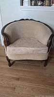Front view of antique single arm chair showing carved wooden arms and legs with beige upholstery and patterned cushion