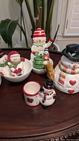 Photo of six holiday-themed ceramic items including a snowman tea pot, snowman figurines, a Christmas tree salt and pepper set, and a small cup on a wooden surface.