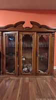 Front view of the antique wooden glass display cabinet showing three glass doors with wood detail, shelves inside with glassware, and the wooden crown molding.