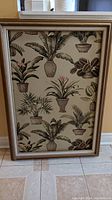 Full view of rectangular canvas picture depicting multiple potted palm trees and tropical plants in a repeated pattern style mounted in a wide gold-toned decorative frame.