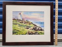 Frontal photo of the framed watercolour painting showing the lighthouse coastal scene with ruler placed nearby for size reference.