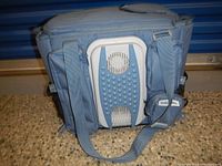 Front view of the cooler travel bag showing blue fabric with dual carrying straps and the thermoelectric cooling unit with fan vents on plastic housing.