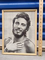 Framed black and white photo print of Bruce Springsteen, approx 13x10 inches, showing him shirtless with a hand near chest, in wood-colored frame.