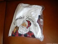 White Santa-themed pajama top folded inside original sealed plastic bag showing the 'Santa's Favorite' graphic with Santa holding a cookie.