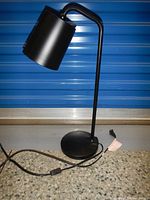 Front view of the black office desk lamp showing the cylindrical shade, slim neck, circular base, and power cord with plug.