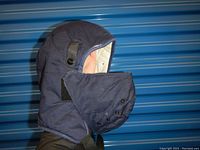 Front angled view of blue cotton winter hat liner showing outer shell, face cover, and straps worn on a head form against a blue background.