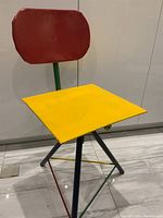 Front view showing the yellow square seat, red rectangular backrest, and multicolored metal tripod base with adjustable height feature.