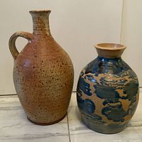 Two vintage Mid-Century Modern studio pottery vases displayed side by side on floor. One vase is tan with speckles and handle, the other is round with blue and tan glaze.