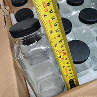 Close-up image showing one glass bottle approximately 7 inches tall with black screw-on cap and measuring tape indicating height.
