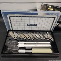 Opened box showing three craft cutter handles, fourteen different shaped blades, and the cutting mat inside a plastic case.