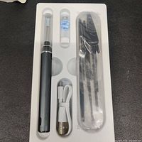 Photo of all components in white molded packaging showing black pen-style ear endoscope, USB cable, multiple black cleaning tools sealed in plastic, and a small container of blue replacement ear picks.