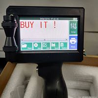 Close-up photo of the handheld printer's touchscreen display showing 'BUY IT!' text and various menu options like Setup, Open, Add, Edit, Save, Delete, and Print.
