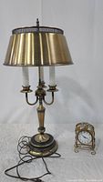 Full view of brass bouilotte lamp with three candles and cord visible, next to vintage metal bird house style clock on a white surface
