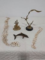 All five items displayed on white quilted surface: seashell plant holder, brass duck, brass seagull, brass dolphin, seashell necklace.