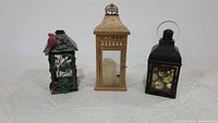 View of three assorted metal lanterns including a rustic wooden-style lantern with candle, a green holly lantern, and a black lantern with Christmas balls.