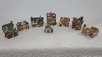 Eight Dept 56 Christmas village houses arranged in a row on a white quilted surface showing variation in roof colors and structure style.