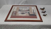 Top-down photo showing the framed folk art print by Paul Jamieson and three duck figurines placed to the right of the frame.
