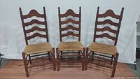 Three Ethan Allen ladderback chairs with rush seats, heirloom nutmeg maple finish, lined up against white background.