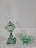Full view of the green Depression glass oil lamp and the Dugan green opalescent fan footed and handled olive dish against a white background.