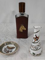 Liquor bottle with stitched brown leather cover and gold horse head motif, Weatherby Hanley Falcon Ware plate with two horse heads and green gold trim, Staffordshire pitcher with hunting scene on white ceramic